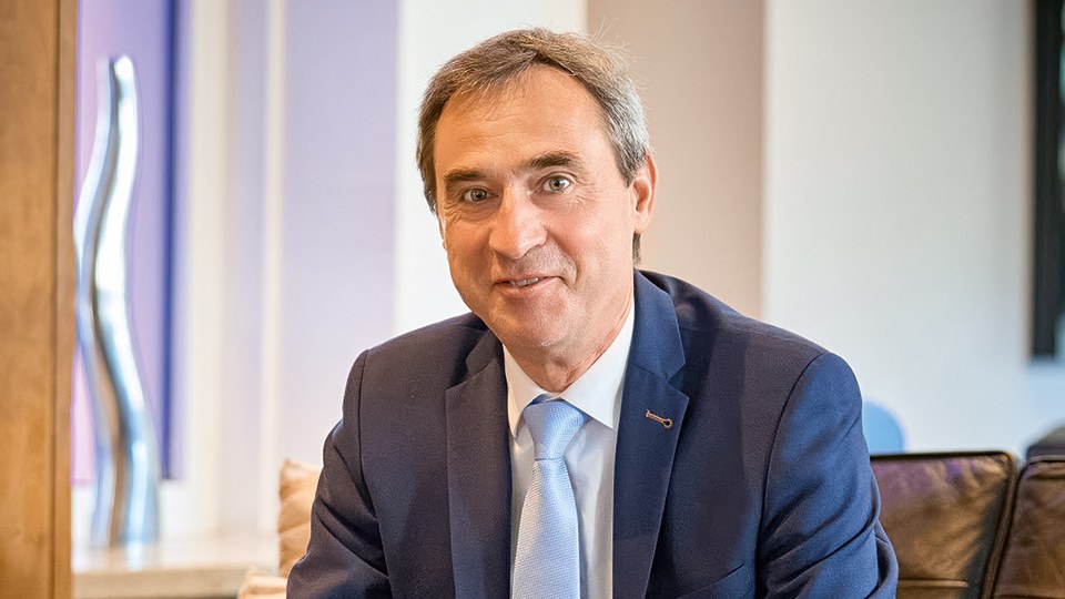 Frank Göller, Leiter der Präventionsabteilung der BG ETEM, sitzt in lockerer Pose auf einem Sofa. Er lehnt sich nach vorn und blickt in die Kamera. Vor ihm ein Glastisch, im Hintergrund unscharf ein Besprechungsraum.