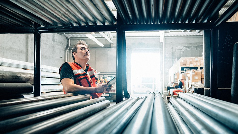 Arbeiter mit Sicherheitsweste, der in einer Lagerhalle einen prüfenden Blick auf ein Regal wirft, in dem lange Metallstangen oder Metallrohre gelagert sind.