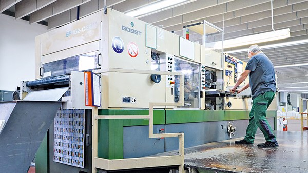 In einer Fabrikhalle steht ein Arbeiter an einer Stanzanlage und zieht mit beiden Händen einen Ausbrechrahmen heraus.