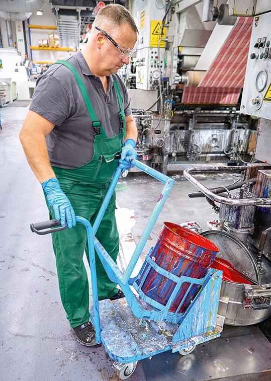Arbeiter mit grüner Arbeitslatzhose, Schutzhandschuhen und Schutzbrille füllt aus einem Farbeimer Farbe an einer Druckmaschine nach. Der Farbeimer steht auf einem Handwagen und wird mithilfe eines Mechanismus gekippt.
