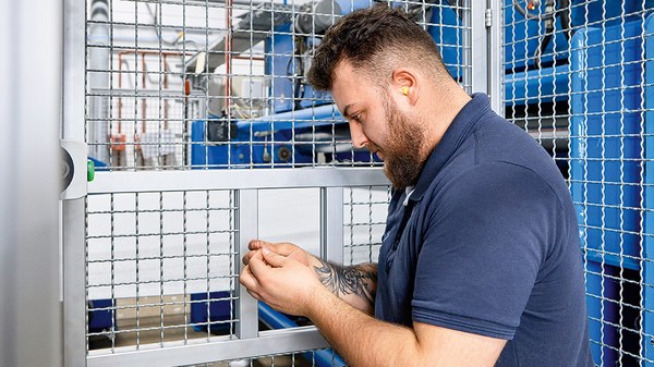 Arbeiter mit blauem Polohemd steht an einer Maschine. Diese ist durch eine Art Käfig geschützt, der Zugriff in ihren Bereich ist nur über einen kleinen fensterähnlichen  Ausschnitt des Käfigs möglich.