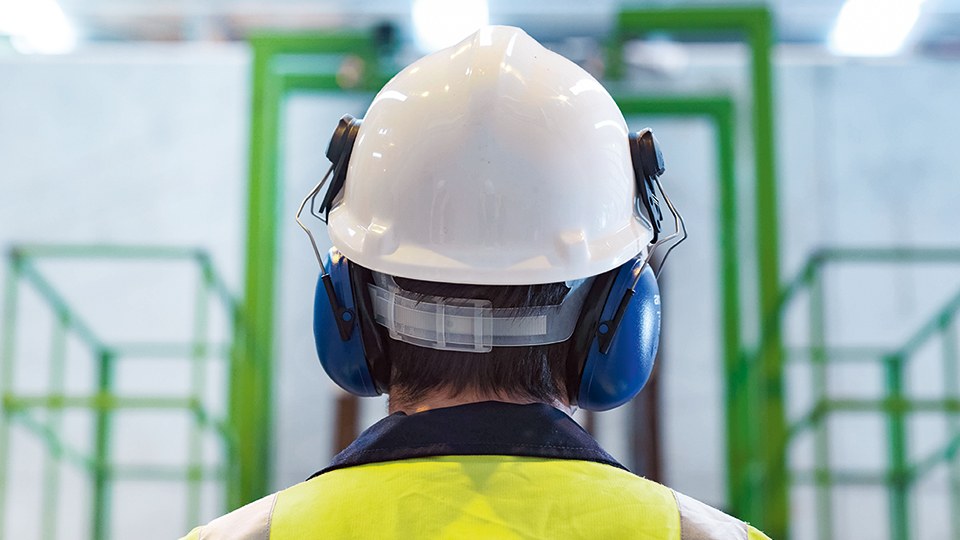 Rückansicht eines Arbeiters in Fabrikhalle. Er trägt Sicherheitsweste, weißen Sicherheitshelm und blaue Kapselgehörschützer.