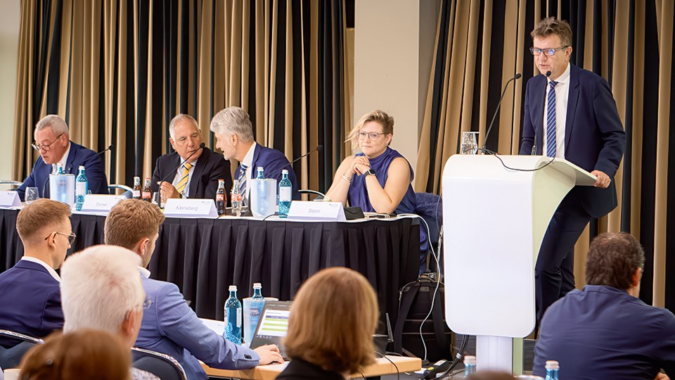 Blick auf das Podium der Vertreterversammlung: Hans-Peter Kern, Franz Donner (Vorsitzende des Vorstands), Jobst Kleineberg, Cathrin Praße-Stern (Vorsitzende der Vertreterversammlung) und Jörg Botti (Hauptgeschäftsführer BG ETEM), der an einem Rednerpult zu den Anwesenden spricht. Im Vordergrund einiger Teilnehmerinnen und Teilnehmer.
