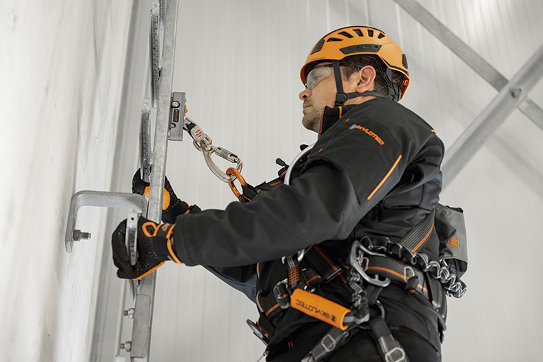Mann in persönlicher Schutzausrüstung in schwarz und orange mit orangenem Helm und Sicherheitsbrille hält sich mit behandschuhten Händen an einer vertikalen Leiter fest, er trägt ein Gurtsystem am Oberkörper und ist mit einem Steigschutzläufer an einer Schiene gesichert.