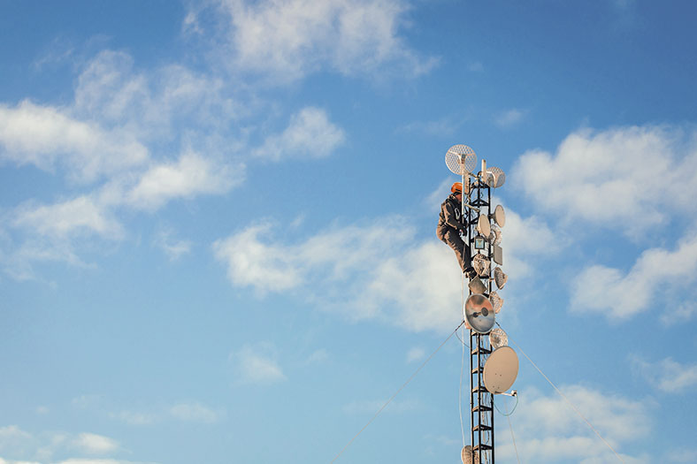 Arbeiter in Schutzausrüstung mit Helm und Gurtsicherung arbeitet an der Spitze eines Antennenträgers mit Satellitenschüsseln, im Hintergrund blauer Himmel mit einigen weißen Wolken.