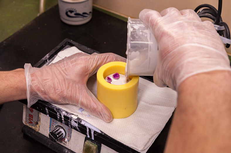 Eine Hand in Gummihandschuhen hält eine gelbe zylindrische Form, die auf einem kleinen Heizgerät steht, während die andere Hand aus einem Plastikbehälter eine weiße Masse hineingießt.