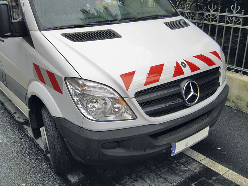 Ausschnitt der Vorderseite eine weißen Mercedes-Transporters mit rot-weißen Warnmarkierungen auf der Motorhaube und am vorderen Kotflügel. Er parkt auf einer Straße neben einem gusseisernen Zaun.