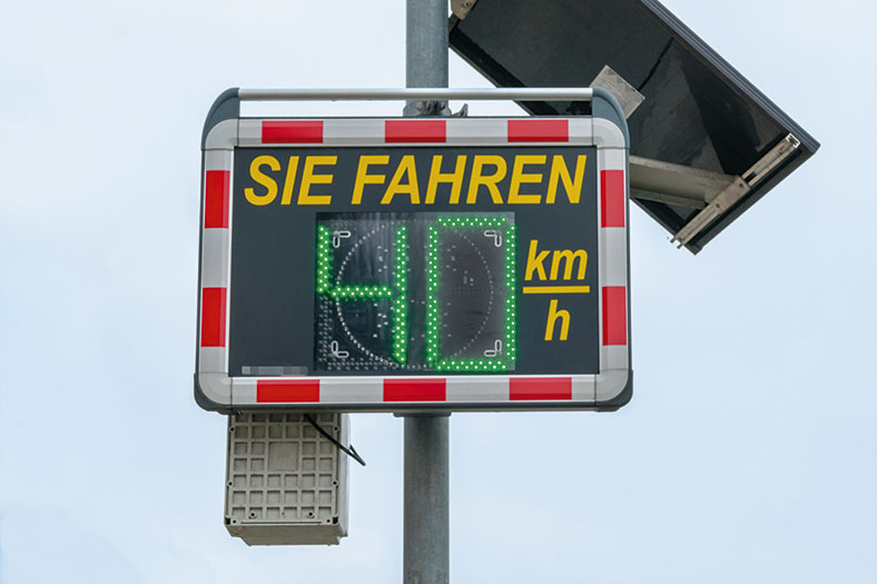 Das Foto zeigt ein solarbetriebene Geschwindigkeitsmass- und Anzeigesystem, das die gefahrene Geschwindigkeit eines sich nähernden Fahrzeugs in grünen Ziffern anzeigt. Die Anzeigetafel hat einen rot-weiß-gestreiften Rand.