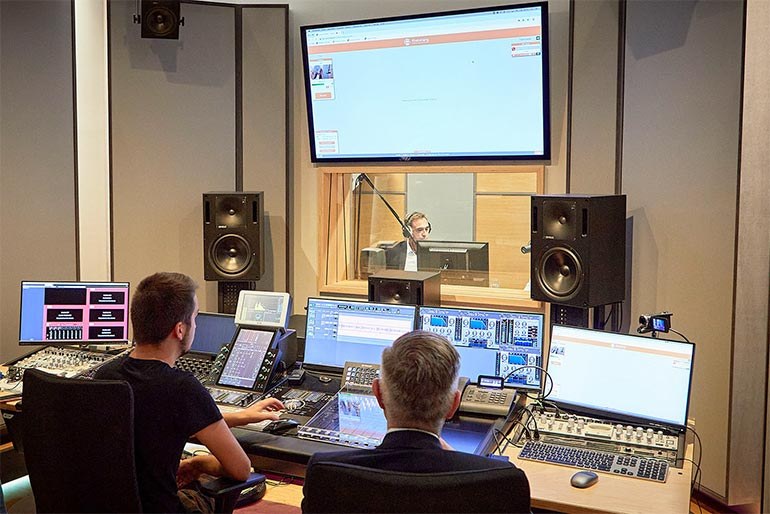 Studioaufnahme für Podcast „Ganz sicher“: Studiotechniker und Beisitzer vor vielen Geräten schauen durch eine Glasscheibe ins Studio, wo ein Mann mit Kopfhörer sitzt.