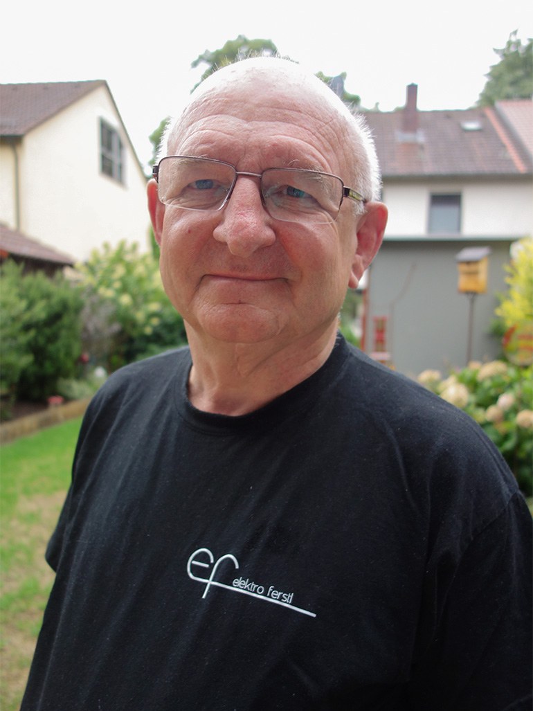 Porträt von Karl Heinz Ferstl, Fa. Elektro Ferstl. Er hat graue Haare, trägt eine Brille und ein schwarzes T-Shirt mit Firmenlogo und steht im Freien.