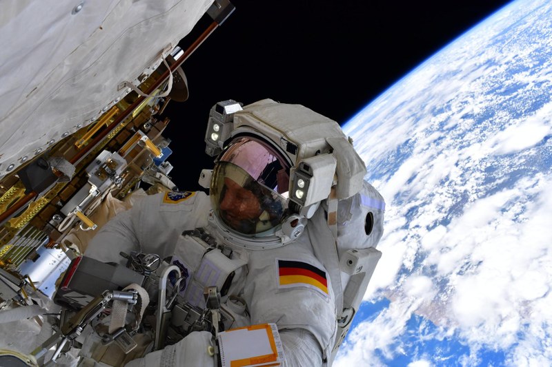 Ein Astronaut schwebt im All neben einer Luftschleuse, im Hintergrund ist die Erde zu sehen.