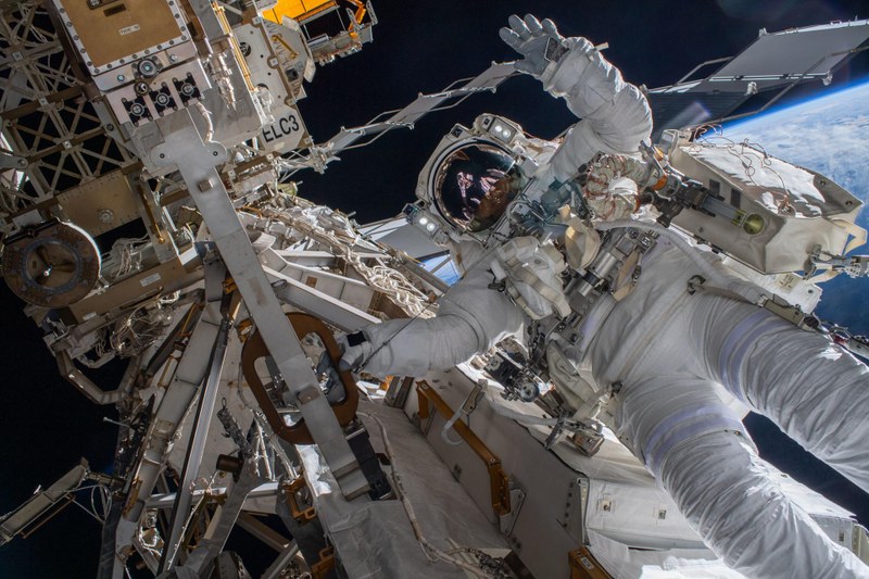 Astronaut winkt auf Außenmission an der ISS-Raumstation, im Hintergrund sieht man den einen Ausschnitt der Erde.
