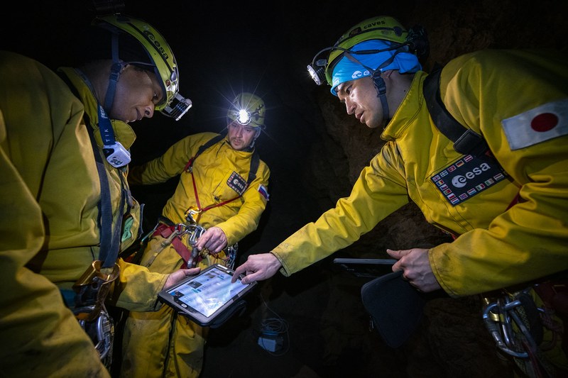 In einer Höhle stehen drei Personen in gelber Schutzkleidung mit blauen Helmen zusammen und schauen auf ein Tablet.