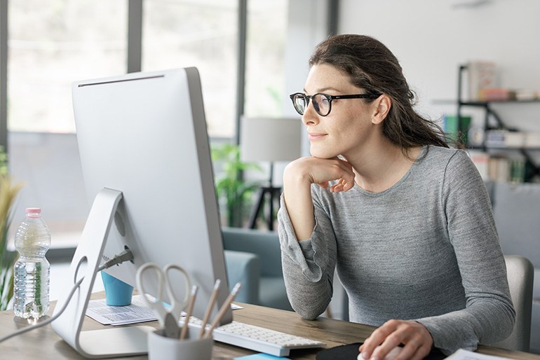Büroszene mit einer jungen Frau in grauem Oberteil mit Brille, die an einem Schreibtisch sitzt und lächelnd auf einen Bildschirm schaut. Von links oben ragt ein Foto des Eifelturms schräg  ins Bild.