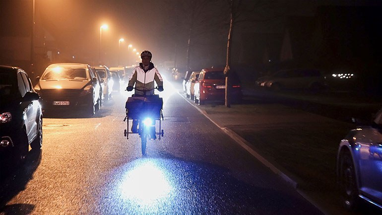 Zeitungszustellung: Zusteller fährt im Dunkeln mit Fahrrad auf einer Straße.