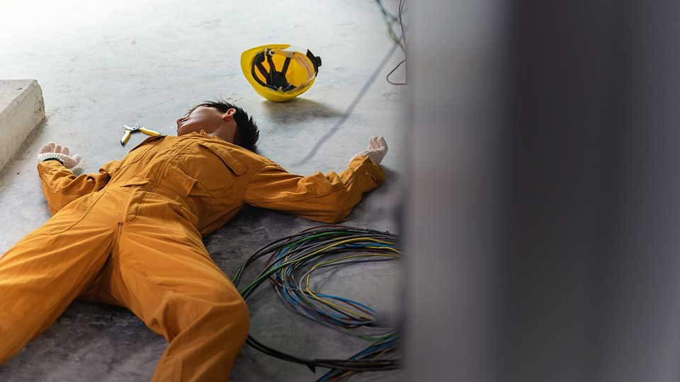 Ein Arbeiter liegt bewusstlos auf dem Boden. Unter und neben ihm Stromkabel und Klemmen.