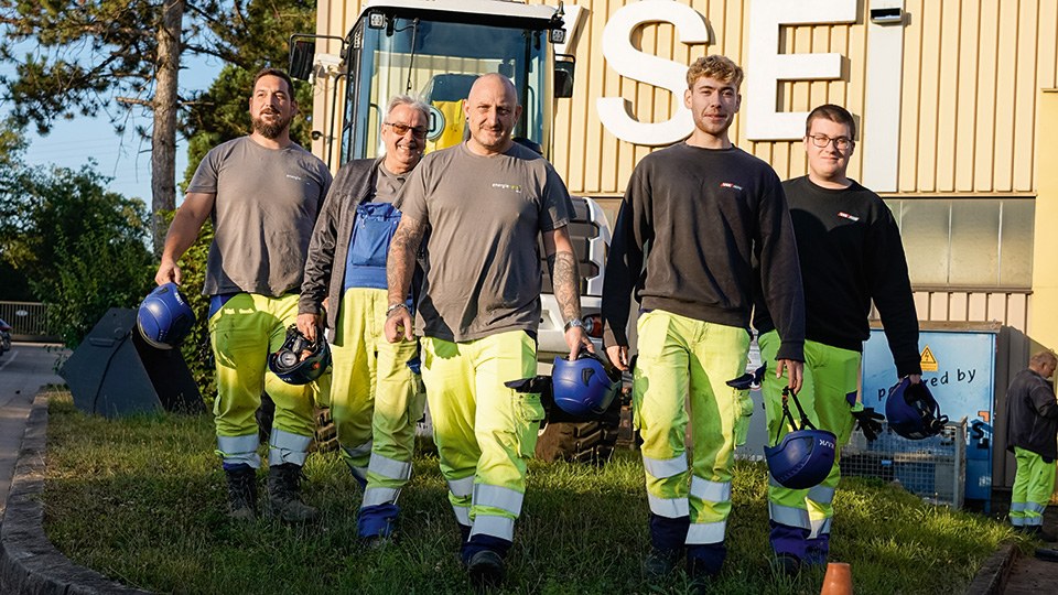 5 Mitarbeiter der VSE-Gruppe laufen auf die Kamera zu; sie tragen reflektierende Sicherheitshosen und halten jeweils einen Sicherheitshelm in der Hand; hinter ihnen ein Bagger und eine Werkshalle, vor ihnen ein Pylon