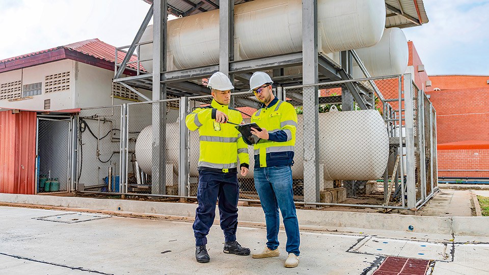 Zwei Arbeiter mit Helm und Warnjacken besprechen sich vor einer Anlage mit großen Gastanks
